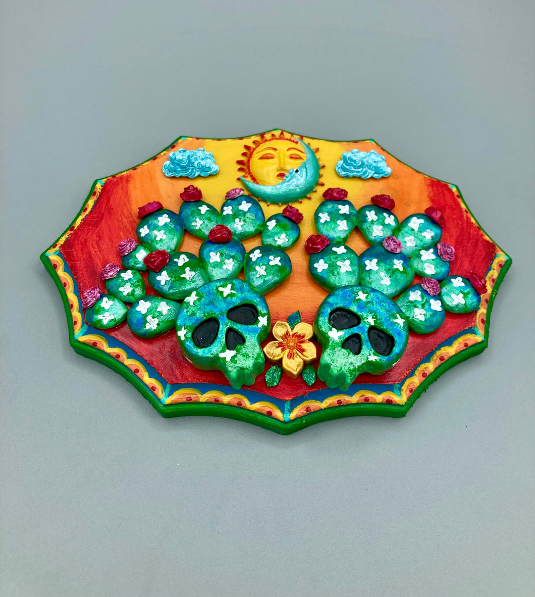 This handcrafted polymer clay wall art features a wood frame painted in a gradient from light yellow to dark red, creating a stunning sunset look. With two bright green skull cactus plant and bright dark red blooming flowers. Featuring a vivid sun and crescent moon with a backdrop of light blue clouds, this frame is sure to make an impression. A bight green hue frame to make a bold contrast with the warm background hue. This piece is sure to make a statement.