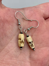 Load image into Gallery viewer, This set of earrings features mini bone-carved skull beads set in black glass. Perfect for goth, emo, witchy or Day of the Dead ensembles, these earrings will bring a unique flair to any outfit.   Measurements: 1 ” in length  
