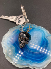 Load image into Gallery viewer, This beautiful edgy gun metal faceted shimmery multi-color glass skull bead keychain is full of hues of silver, purple, pink, blue, and gold. The split nickel key ring is 1 1/4&quot; in diameter and the entire key chain is 4 1/2&quot; in length (from key ring to skull bead)  Measurements: Nickel Free Key Ring is 1 1/4” in diameter, Entire Key Chain is 4 1/2&quot; in Length, Metal Skull Bead 5/8&quot; in Height
