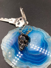 Load image into Gallery viewer, This beautiful edgy gun metal faceted shimmery multi-color glass skull bead keychain is full of hues of silver, purple, pink, blue, and gold. The split nickel key ring is 1 1/4&quot; in diameter and the entire key chain is 4 1/2&quot; in length (from key ring to skull bead)  Measurements: Nickel Free Key Ring is 1 1/4” in diameter, Entire Key Chain is 4 1/2&quot; in Length, Metal Skull Bead 5/8&quot; in Height
