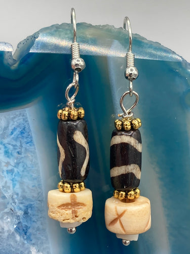 This set of earrings is a one-of-a-kind combination of cylinder and round bone beads with wavy line designs, enhanced with a floral gold tone metal bead set. One earring features vertical wavy lines and crosses, while the other showcases horizontal wavy lines with an 