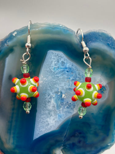 Vibrant, bumpy round lampworked glass beaded earrings in an array of colors including light green, white, yellow, dark red and bright orange. The fiesta piñata-shaped bead invokes a festive vibe, while its unique virus-shaped design promotes a bold statement. An eye-catching and unique piece of jewelry sure to turn heads. Measurements: 1 1/4” in length
