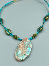 Load image into Gallery viewer, This stunning two-strand necklace features an array of glass beads in hues of green and blue turquoise, gold and green for a vibrant, colorful and bold look. The centerpiece is a large howlite stone chip dyed in turquoise color, accented by the bronze patina cowry beads.  Measurements: Necklace is 17” 
