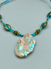 Load image into Gallery viewer, This stunning two-strand necklace features an array of glass beads in hues of green and blue turquoise, gold and green for a vibrant, colorful and bold look. The centerpiece is a large howlite stone chip dyed in turquoise color, accented by the bronze patina cowry beads.  Measurements: Necklace is 17” 
