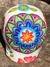 Load image into Gallery viewer, Inspired by nature, this ceramic skull statuette is full of vibrant color and intricate details. The primary color is white with multi-color floral motifs that are painted across the skull, along with hearts and leaves and lively colorful accents around the entire skull. 
