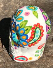 Load image into Gallery viewer, Inspired by nature, this ceramic skull statuette is full of vibrant color and intricate details. The primary color is white with multi-color floral motifs that are painted across the skull, along with hearts and leaves and lively colorful accents around the entire skull. 

