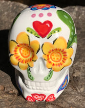 Load image into Gallery viewer, Inspired by nature, this ceramic skull statuette is full of vibrant color and intricate details. The primary color is white with multi-color floral motifs that are painted across the skull, along with hearts and leaves and lively colorful accents around the entire skull.
