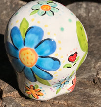 Load image into Gallery viewer, Inspired by nature, this ceramic skull statuette is full of vibrant color and intricate details. The primary color is white with multi-color floral motifs that are painted across the skull, along with hearts and leaves and lively colorful accents around the entire skull. 
