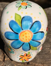 Load image into Gallery viewer, Inspired by nature, this ceramic skull statuette is full of vibrant color and intricate details. The primary color is white with multi-color floral motifs that are painted across the skull, along with hearts and leaves and lively colorful accents around the entire skull. 
