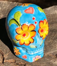 Load image into Gallery viewer, Inspired by nature, this ceramic skull statuette is full of vibrant color and intricate details. The primary color is blue with multi-color floral motifs that are painted across the skull, along with hearts and leaves and lively colorful accents around the entire skull. 
