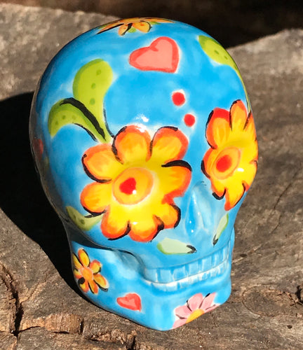 Inspired by nature, this ceramic skull statuette is full of vibrant color and intricate details. The primary color is blue with multi-color floral motifs that are painted across the skull, along with hearts and leaves and lively colorful accents around the entire skull. 