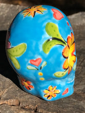 Load image into Gallery viewer, Inspired by nature, this ceramic skull statuette is full of vibrant color and intricate details. The primary color is blue with multi-color floral motifs that are painted across the skull, along with hearts and leaves and lively colorful accents around the entire skull. 

