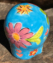 Load image into Gallery viewer, Inspired by nature, this ceramic skull statuette is full of vibrant color and intricate details. The primary color is blue with multi-color floral motifs that are painted across the skull, along with hearts and leaves and lively colorful accents around the entire skull. 
