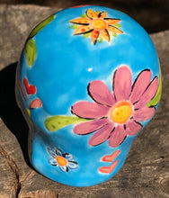 Load image into Gallery viewer, Inspired by nature, this ceramic skull statuette is full of vibrant color and intricate details. The primary color is blue with multi-color floral motifs that are painted across the skull, along with hearts and leaves and lively colorful accents around the entire skull. 
