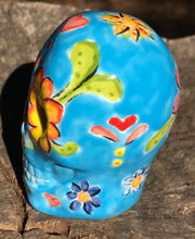 Load image into Gallery viewer, Inspired by nature, this ceramic skull statuette is full of vibrant color and intricate details. The primary color is blue with multi-color floral motifs that are painted across the skull, along with hearts and leaves and lively colorful accents around the entire skull. 

