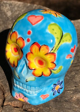 Load image into Gallery viewer, Inspired by nature, this ceramic skull statuette is full of vibrant color and intricate details. The primary color is blue with multi-color floral motifs that are painted across the skull, along with hearts and leaves and lively colorful accents around the entire skull. 
