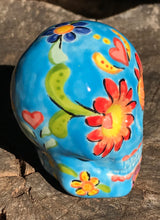 Load image into Gallery viewer, Inspired by nature, this ceramic skull statuette is full of vibrant color and intricate details. The primary color is sky blue with Multi-color floral motifs that are painted across the skull, along with hearts and leaves and lively colorful accents around the entire skull. 
