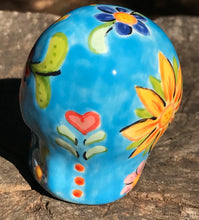 Load image into Gallery viewer, Inspired by nature, this ceramic skull statuette is full of vibrant color and intricate details. The primary color is sky blue with Multi-color floral motifs that are painted across the skull, along with hearts and leaves and lively colorful accents around the entire skull. 
