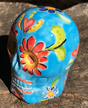 Load image into Gallery viewer, Inspired by nature, this ceramic skull statuette is full of vibrant color and intricate details. The primary color is sky blue with Multi-color floral motifs that are painted across the skull, along with hearts and leaves and lively colorful accents around the entire skull. 
