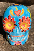 Load image into Gallery viewer, Inspired by nature, this ceramic skull statuette is full of vibrant color and intricate details. The primary color is sky blue with Multi-color floral motifs that are painted across the skull, along with hearts and leaves and lively colorful accents around the entire skull. 
