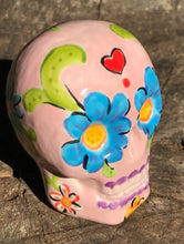 Load image into Gallery viewer, Inspired by nature, this ceramic skull statuette is full of vibrant color and intricate details. The primary color is soft pink with multi-color floral motifs that are painted across the skull, along with hearts and leaves and lively colorful accents around the entire skull. 

