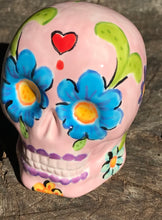 Load image into Gallery viewer, Inspired by nature, this ceramic skull statuette is full of vibrant color and intricate details. The primary color is soft pink with multi-color floral motifs that are painted across the skull, along with hearts and leaves and lively colorful accents around the entire skull. 

