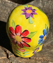 Load image into Gallery viewer, Inspired by nature, this ceramic skull statuette is full of vibrant color and intricate details. The primary color is yellow with Multi-color floral motifs that are painted across the skull, along with hearts and leaves and lively colorful accents around the entire skull.
