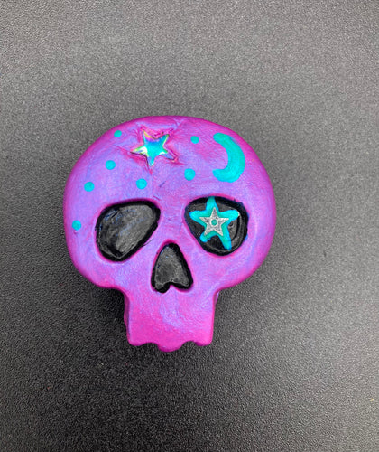 Cute and spooky magnet design to adorn your refrigerator, lockers, workstation, toolbox, or dishwasher. This skull statuette is a tribute to nature. Its metallic violet hue evokes sundown, transitioning the sky from blue to purple to black. Adding to the appeal of this piece is a teal-green moon and stars that provide a dramatic contrast against the violet. The forehead is finished with a rainbow hematite star bead for a unique accent.  H 1 1/2” x W 1 1/2” x D 3/4”