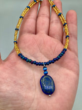 Load image into Gallery viewer, This Lapis pendant necklace features dark blue cats eye beads and two strands of gold glass beads with dark blue double hole beads. The large multi-colored glass beads provide remarkable shimmer and a full range of colors, such as purples, blues, greens, and golds, that enhances the lapis stone pendant.   Measurements:   Necklace is 19” in length 
