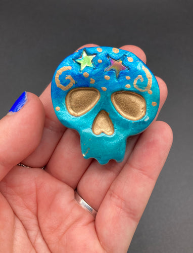 Perfect item to add to your fridge, locker, cubicle, toolbox or dishwasher. This handcrafted skull statuette features shimmering metallic and vivid blue hues, metallic gold stars, and intricate detailing. The skull's forehead is adorned with a glittering hematite star bead.  Measurements: H 1 1/2” x W 1 1/2” x D 3/4” 