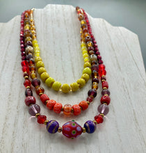 Load image into Gallery viewer, This necklace consists of three strands with a multitude of shades ranging from red to orange, gold, and amber, providing a captivating visual combination. Crafted with a combination of glass, ceramic and metal beads, this eye-catching piece of jewelry radiates power and strength.  Measurements:  Necklace is 16” in length, can be adjusted up to 17 1/2&quot;
