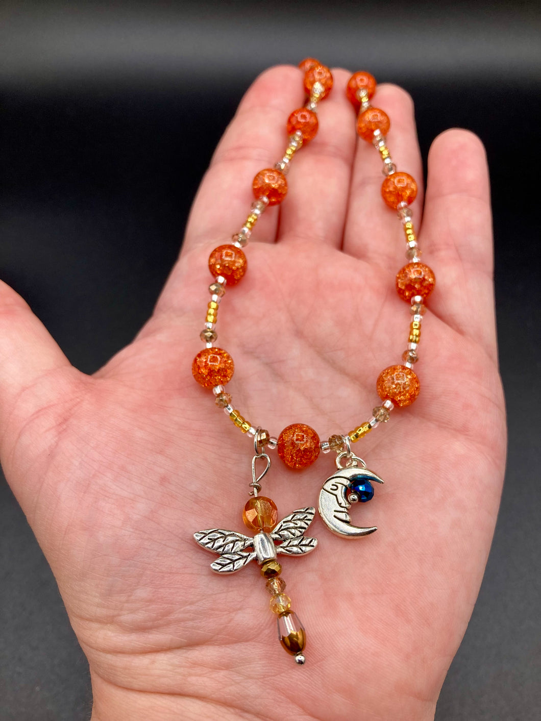 This bright orange, adjustable, choker-style necklace is adorned with beaded glass and a striking crescent moon charm and dragonfly pendant. Accessorize with matching dragonfly earrings for an eye-catching, complementary look that captures the warmth of summer. Perfect for any outfit, its vibrant colors will make a statement and complete any look.  Measurements: Necklace is 16 1/2”adjustable up to 19