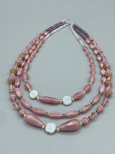 This three-strand beaded necklace is crafted with iridescent dusty rose glass, along with white, gold, pink, and iridescent purple beads for an eclectic mix of colors, tones, and textures. Perfect for any occasion, this gorgeous statement piece will add the finishing touch to your look.   Measurements:   Necklace is 17” in length, can be adjusted up to 19