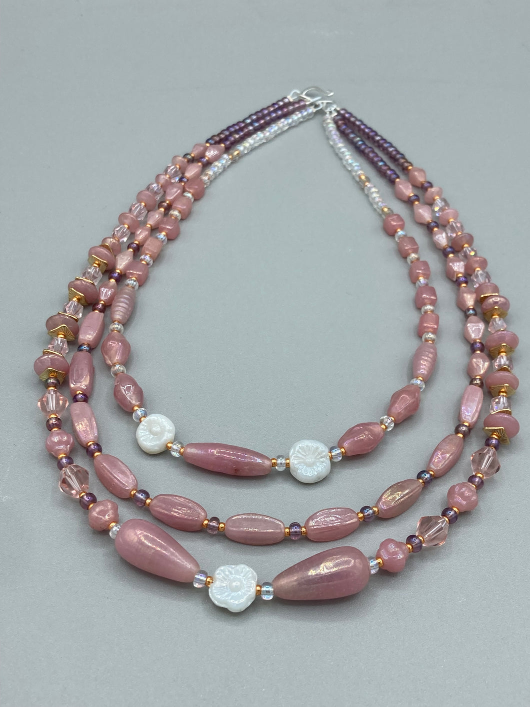 This three-strand beaded necklace is crafted with iridescent dusty rose glass, along with white, gold, pink, and iridescent purple beads for an eclectic mix of colors, tones, and textures. Perfect for any occasion, this gorgeous statement piece will add the finishing touch to your look.   Measurements:   Necklace is 17” in length, can be adjusted up to 19