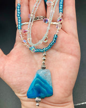 Load image into Gallery viewer, This exquisite necklace features a vibrant Sea Blue Agate Natural Stone Pendant and two strands of multi-colored glass beaded necklace. An adjustable collar-style strand contains small, light blue beads and lots of iridescent double-hole beads for shimmer and sparkle. The longer strand features larger blue glass beads interspersed with clear and silver beads for a sophisticated, draped look. It's finished with a set of dainty matching earrings for a complete look. Necklace is 14”adjustable up to 18"

