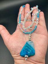 Load image into Gallery viewer, This exquisite necklace features a vibrant Sea Blue Agate Natural Stone Pendant and two strands of multi-colored glass beaded necklace. An adjustable collar-style strand contains small, light blue beads and lots of iridescent double-hole beads for shimmer and sparkle. The longer strand features larger blue glass beads interspersed with clear and silver beads for a sophisticated, draped look. It's finished with a set of dainty matching earrings for a complete look. Necklace is 14”adjustable up to 18"
