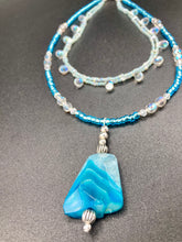 Load image into Gallery viewer, This exquisite necklace features a vibrant Sea Blue Agate Natural Stone Pendant and two strands of multi-colored glass beaded necklace. An adjustable collar-style strand contains small, light blue beads and lots of iridescent double-hole beads for shimmer and sparkle. The longer strand features larger blue glass beads interspersed with clear and silver beads for a sophisticated, draped look. It's finished with a set of dainty matching earrings for a complete look. Necklace is 14”adjustable up to 18"
