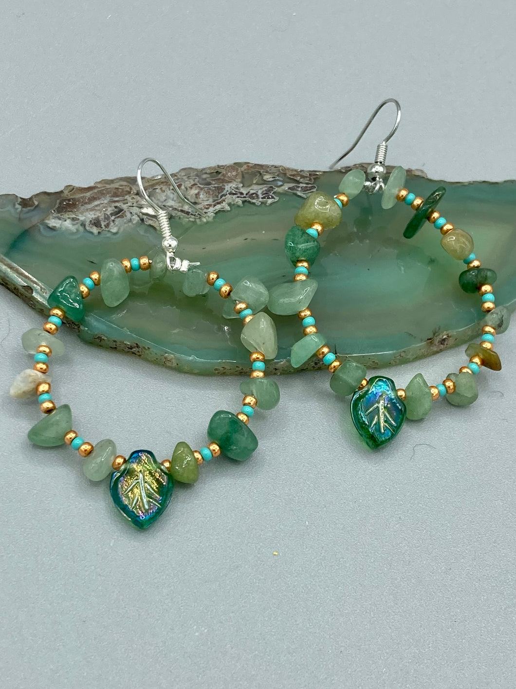 Natural aventurine stone chips, turquoise, and gold glass beads are strung together to form these stylish hoop earrings. The dark green pressed glass leaf charm complements the design, creating an iridescent, shimmery finish with subtle hues of gold, purple, blue, and pink. Measurements: 2