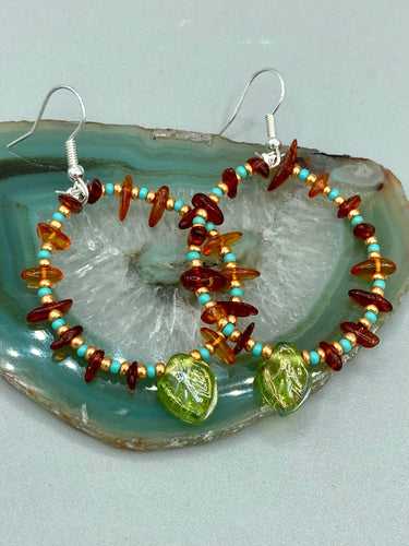 These Amber Stone Chip Beaded Hoop Earrings feature turquoise and gold glass beads, and a light green glass leaf charm with an iridescent shimmer finish with hints of gold, purple, blue, and pink.  Measurements: 2