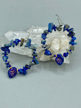 Load image into Gallery viewer, These Lapis Stone Chip Beaded Hoop Earrings feature a dark blue-tone pressed glass leaf charm with an iridescent shimmer finish showcasing hues of gold, purple, blue and pink. The earrings display natural lapis stone chips and royal blue with gold glass beads.  Measurements: 1 1/2&quot; length
