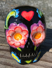 Load image into Gallery viewer, Inspired by nature, this ceramic skull statuette is full of vibrant color and intricate details. The primary color is black with multi-color floral motifs that are painted across the skull, along with hearts and leaves and lively colorful accents around the entire skull. 
