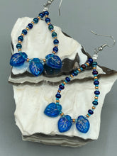 Load image into Gallery viewer, These striking Royal Blue Pressed Glass Leaf Beaded Hoop Earrings feature a shimmery finish with a hint of gold, purple, blue and pink. Gold, dark and light blue glass beads complete the piece.  Measurements: 2 1/2&quot; length
