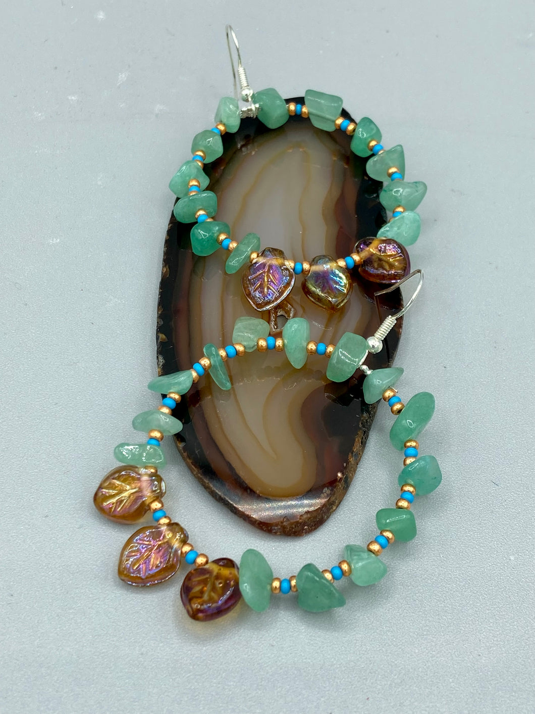 These Aventurine Stone Chip Beaded Hoop Earrings feature natural aventurine stone chips, turquoise, and gold glass beads. The amber-tone pressed glass leaf charm has an iridescent shimmer finish, with subtle accents of gold, purple, blue, and pink. Measurements: 2