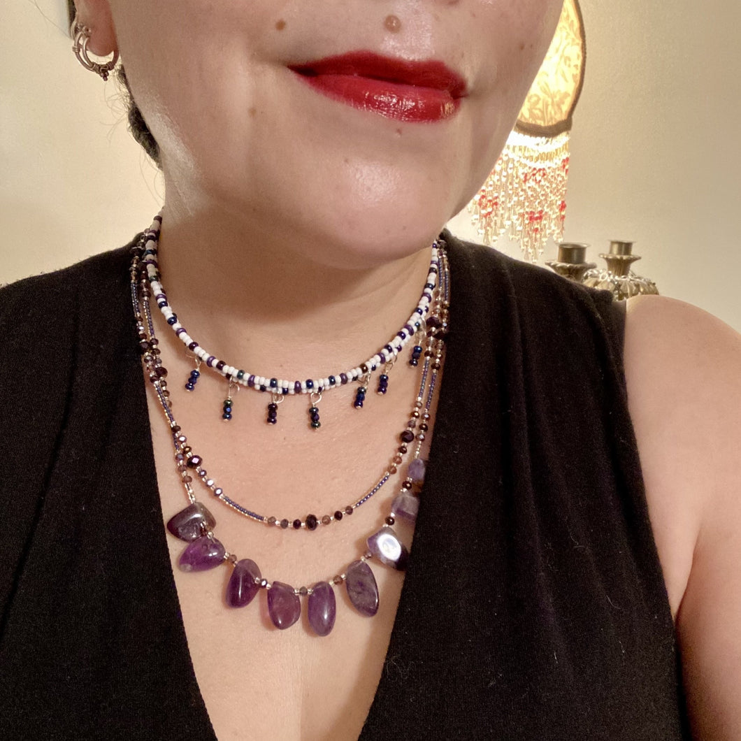 The Genuine Amethyst Natural Gemstone Adjustable Chocker Style Glass Beaded Necklace features vibrant violet and white tones. Its unique draped beading creates a beautiful style with a series of short strands of glass beads for movement and a longer strand with a multitude of amethyst nugget beads. The collar necklace hangs gracefully along the neck and chest, making it a dream come true for violet lovers.  Measurements:  Necklace is 15” in length adjustable to 18