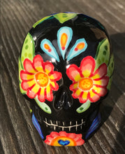 Load image into Gallery viewer, Inspired by nature, this ceramic skull statuette is full of vibrant color and intricate details. The primary color is black with multi-color floral motifs that are painted across the skull, along with hearts and leaves and lively colorful accents around the entire skull. 

