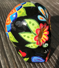 Load image into Gallery viewer, Inspired by nature, this ceramic skull statuette is full of vibrant color and intricate details. The primary color is black with multi-color floral motifs that are painted across the skull, along with hearts and leaves and lively colorful accents around the entire skull. 
