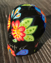 Load image into Gallery viewer, Inspired by nature, this ceramic skull statuette is full of vibrant color and intricate details. The primary color is black with multi-color floral motifs that are painted across the skull, along with hearts and leaves and lively colorful accents around the entire skull. 
