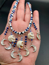 Load image into Gallery viewer, Howlite Skull Beads And Smiling Crescent Moon Charms Choker Style Glass Beaded Necklace. Two beaded stands that are primarily white and added purple glass beads that have hues of blue and gold. I wanted to incorporate dark and white glass bead colors to create a simple contrast. Large amount of dangling skulls and their smiling crescent moons add a lot of movement. Necklace is 16”adjustable up to 18" in length - hanging howlite skull with crescent moons are 2” Height
