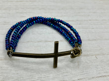 Load image into Gallery viewer, For Positivity and Faith, Shimmery Blue Glass Beaded Bracelet With Curved Horizontal Brass Cross. This bracelet has a large brass cross that is slightly curved to be comfortably worm around your wrist. I created three glass beaded strands that have a wide variety of blue, purple, green, and gold tones. Measurements: 7 1/2&quot; in length, brass cross measures 2&quot; in length
