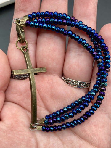 For Positivity and Faith, Shimmery Blue Glass Beaded Bracelet With Curved Horizontal Brass Cross. This bracelet has a large brass cross that is slightly curved to be comfortably worm around your wrist. I created three glass beaded strands that have a wide variety of blue, purple, green, and gold tones. Measurements: 7 1/2
